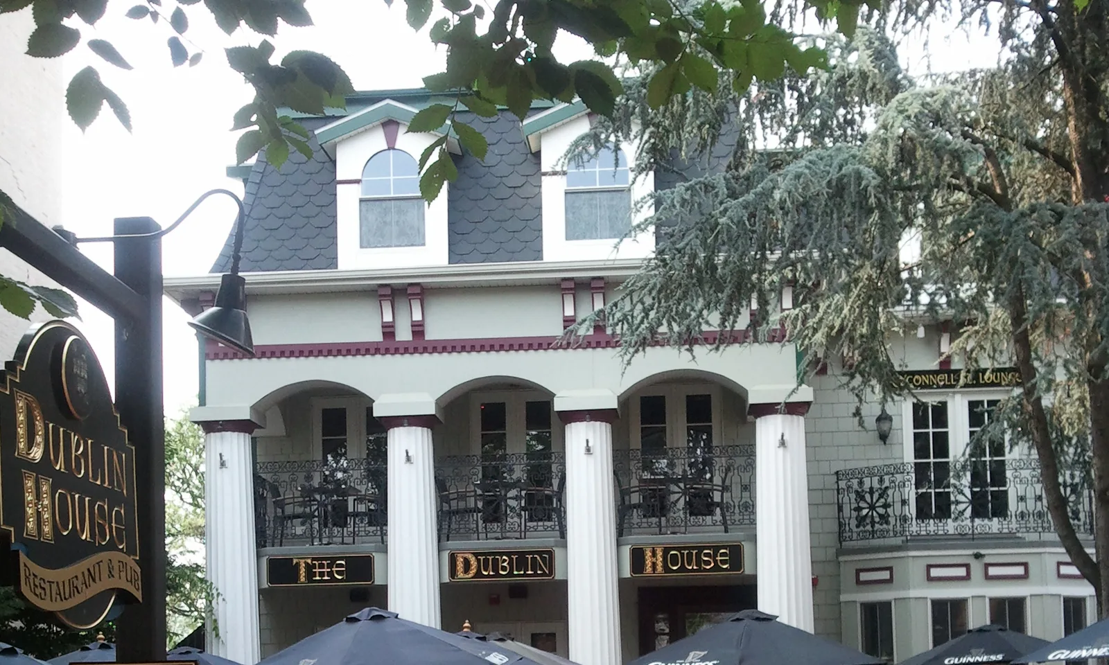 The Dublin House on Monmouth Street: two-storey white columns, gold THE DUBLIN HOUSE boards across the facade, a slate mansard roof over oxblood trim, the carved hanging sign on its bracket, and Guinness umbrellas over the courtyard below